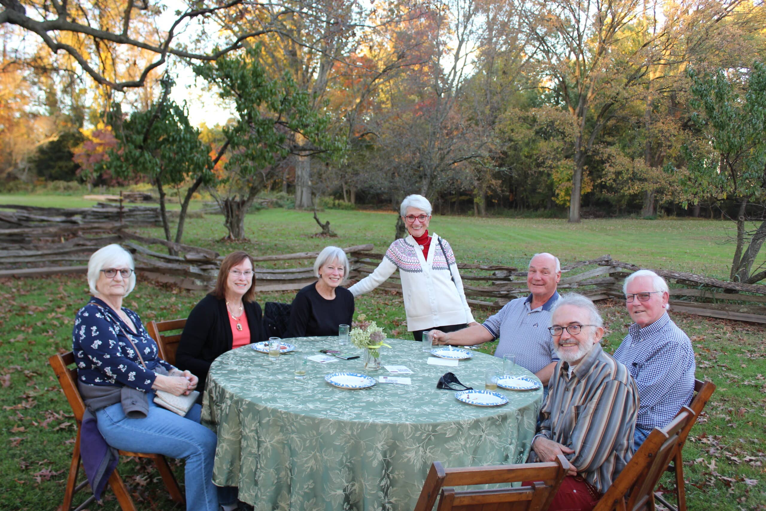 7 people smiling around an outdoors table covered with a table cloth